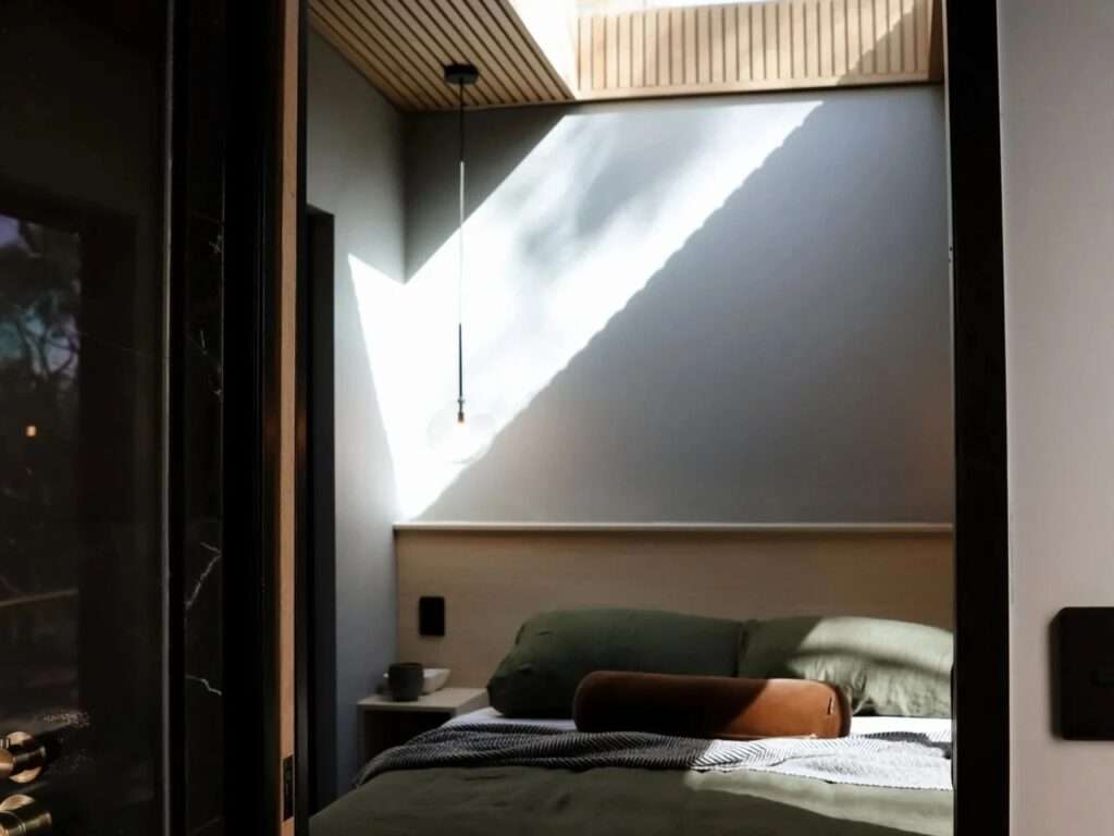 A cozy cabin bedroom viewed through a dark frame, featuring a bed with olive green linen, a large geometric skylight with slatted wood details, and a modern glass pendant light.