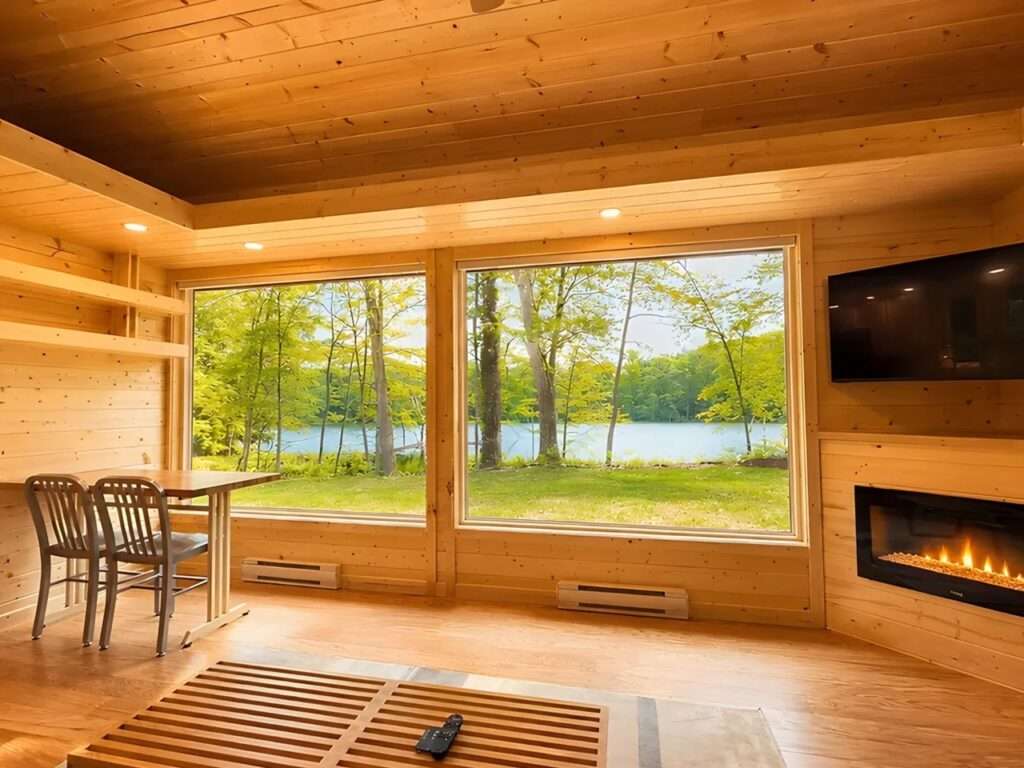 Interior view of a living space with large glass windows framing an outdoor lake view, featuring a wooden dining table with chairs and an integrated electric fireplace on a wood-clad wall.
