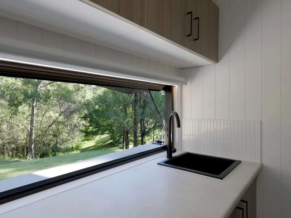 Close-up of a stone kitchen countertop with a matte black faucet, integrated black sink, and an open awning pass-through window looking out to a lush green forest.