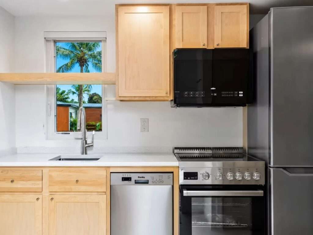 Modern, efficient kitchen interior of the N2 House, featuring light wood cabinets, stainless steel appliances, a range oven, a dishwasher, a microwave, and a window with a view of a palm tree.