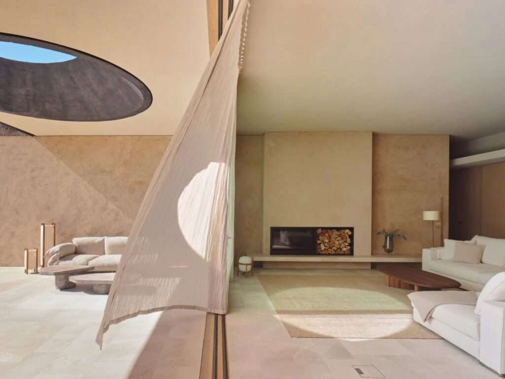 An interior living area with a double fireplace and wood storage, where the sheer curtain is blown by a breeze under a large oculus in the ceiling.