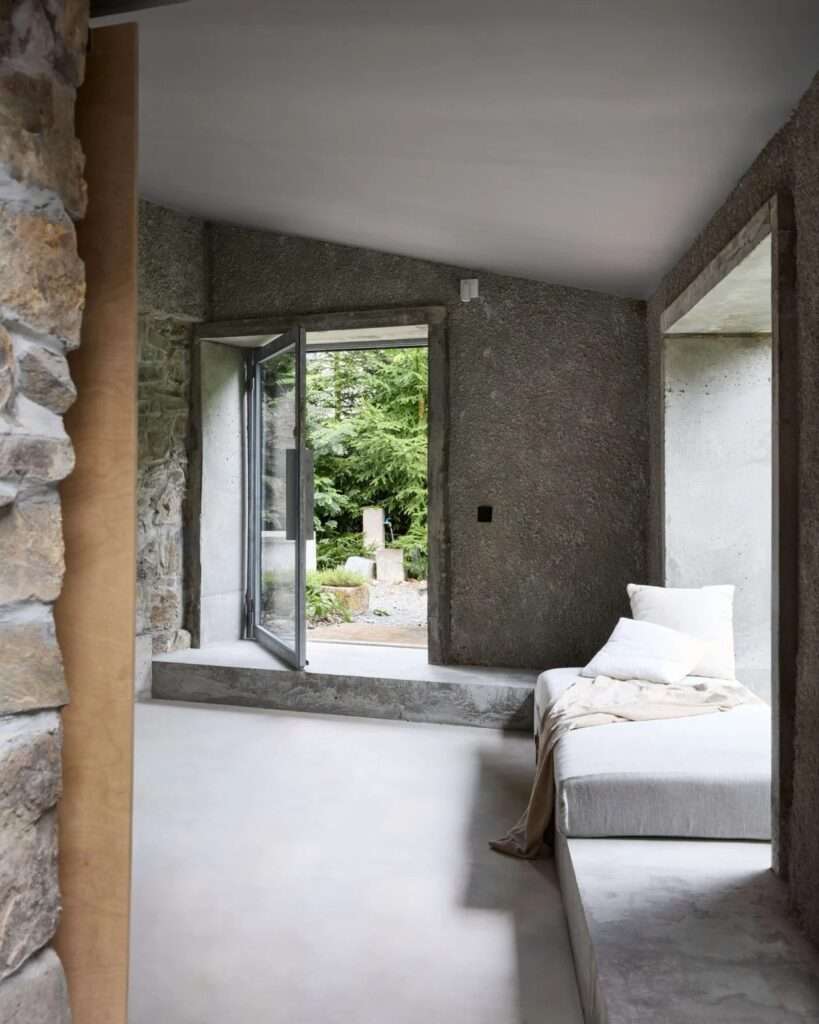 Sunlit living area of adaptive reuse summer house with open glass pivot door, cast-concrete floor, daybed, and exposed stone wall.