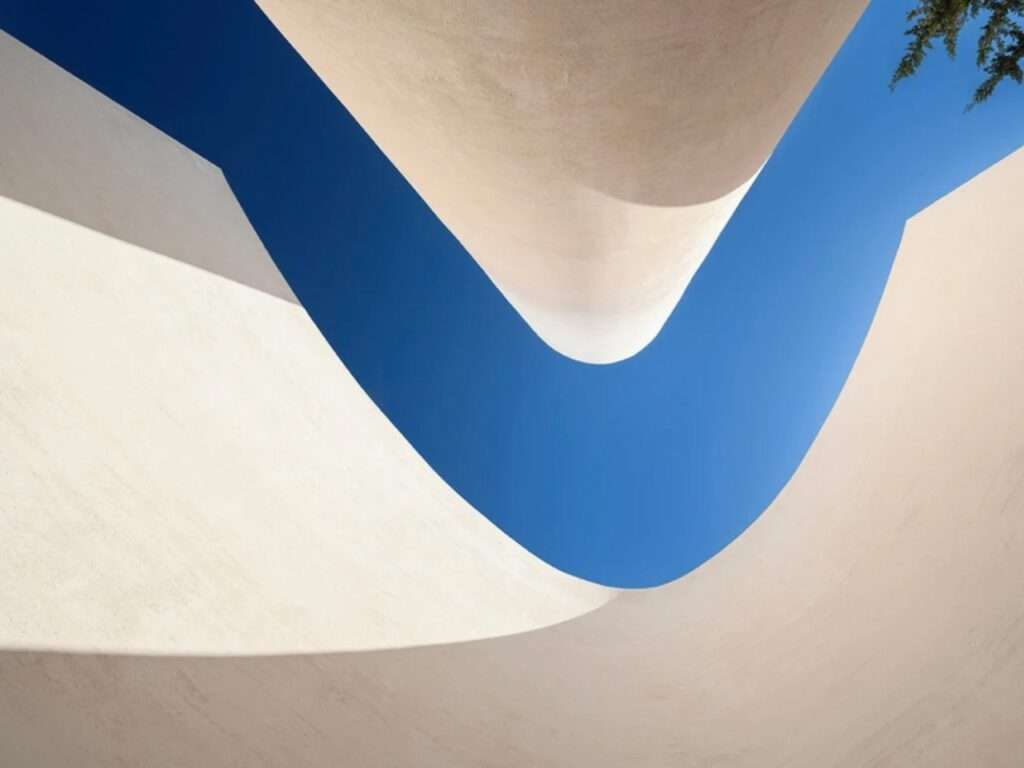 Abstract upward view capturing the curved white concrete wall and the cylindrical bell tower of Santa Barbara Parish against the sky.