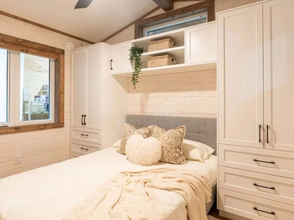 Modern tiny home bedroom with a double bed, gray fabric headboard, built-in floor-to-ceiling white wardrobes, and a high transom window.