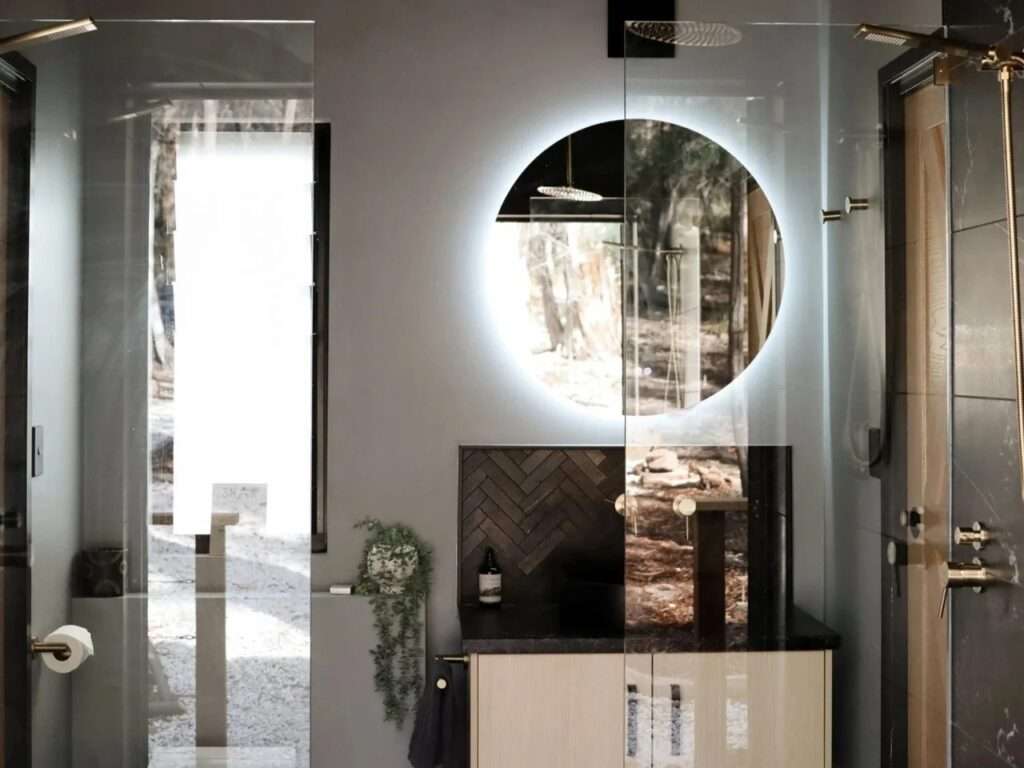Modern cabin bathroom interior with dark tiled walls, a wooden slatted ceiling, a double rain shower with brass fixtures, a floating light wood vanity, and a large round backlit mirror.