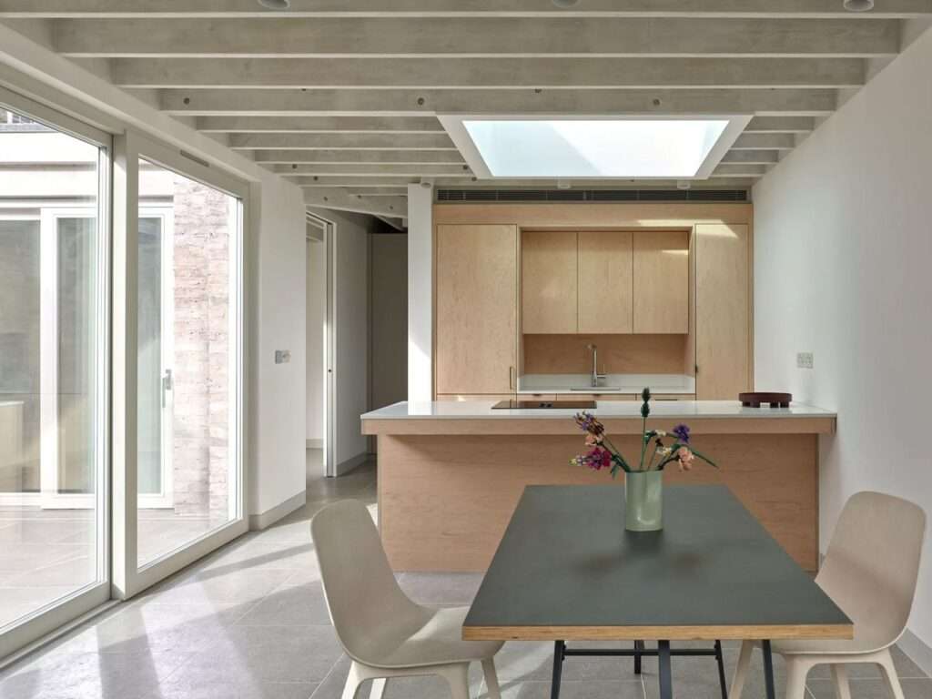 Modern open-plan kitchen and dining area featuring light wood cabinets, white countertops, exposed ceiling beams, and a large central skylight.