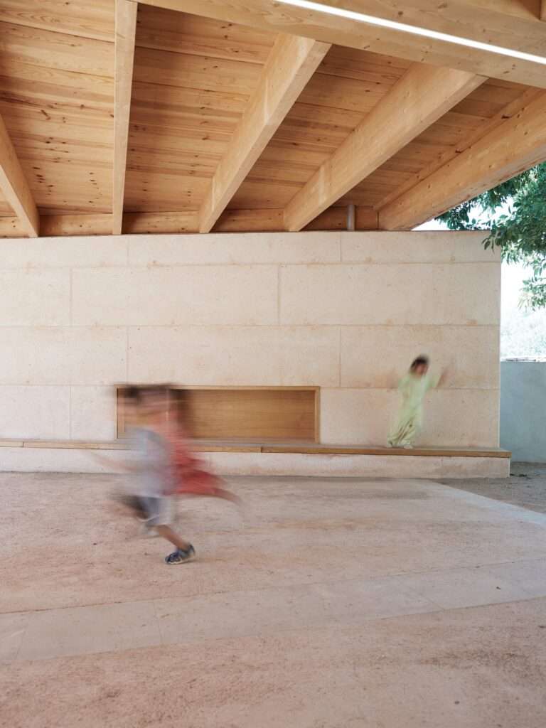 Blurred movement of children running and jumping past a recessed wooden bench integrated into a textured concrete wall.