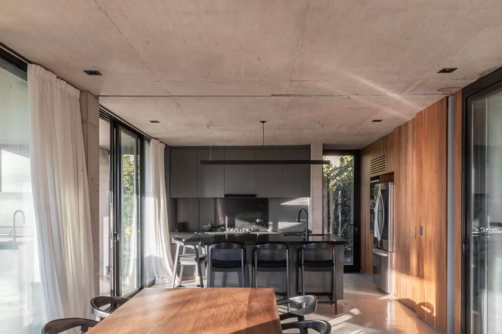 A minimalist kitchen interior with integrated dark matte cabinetry, a central island with bar stools, and dual-aspect glass walls overlooking internal and external gardens.