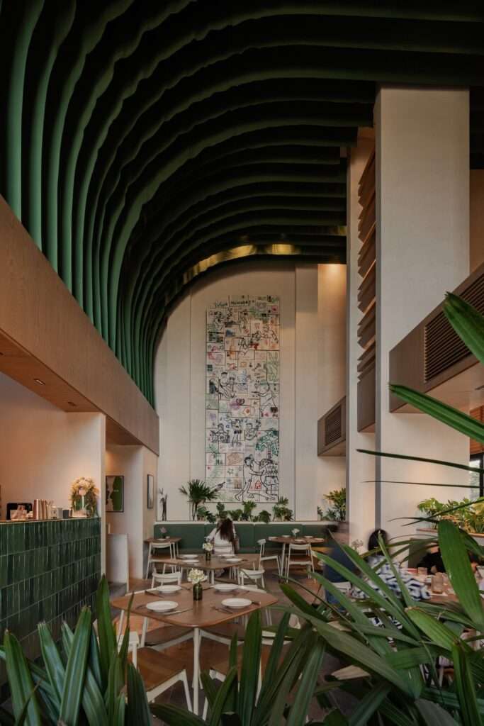 Eye-level view across the main dining area towards a tall gallery wall featuring a vertical recycled book mural framed by green ceiling drapes.