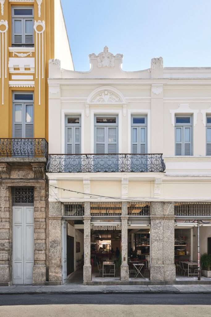 Facade of the Fábrica Bhering adaptive reuse in Rio de Janeiro, Brazil. View from the street showing a two-story building with a light-colored stucco exterior, stone base, large windows, iron railings on a balcony, and decorative architectural elements. Photographed by Rafael Salim.