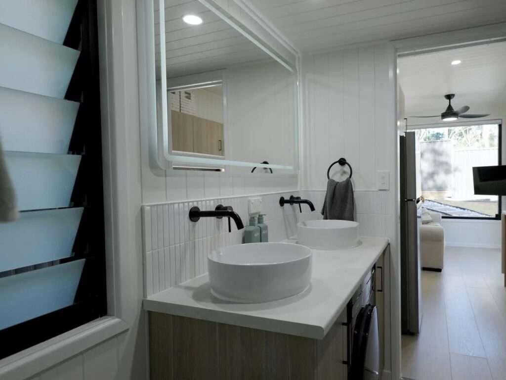 Minimalist bathroom vanity inside the Burley 9.6 house featuring a white stone counter, dual ceramic vessel sinks, matte black wall-mounted faucets, and a large backlit mirror.