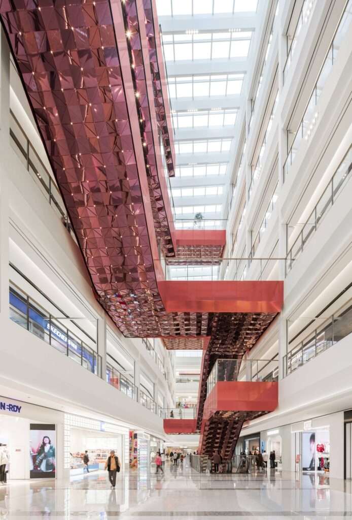 Vertical view of interwoven red escalators and bridges crossing the open-air atrium at Wuxi MixC Mall, with a glossy polished stone floor reflecting the structures.