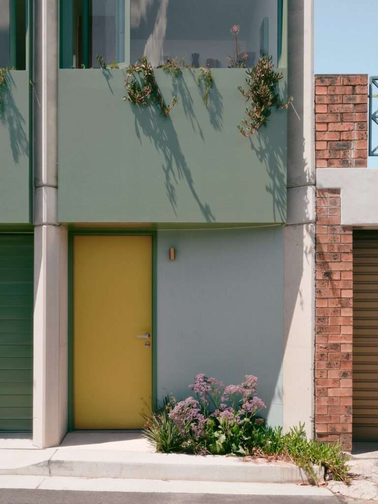 Architectural close-up of a bright yellow entry door adjacent to a green garage roller door, framed by concrete pillars, light blue tiles, and purple flowers.