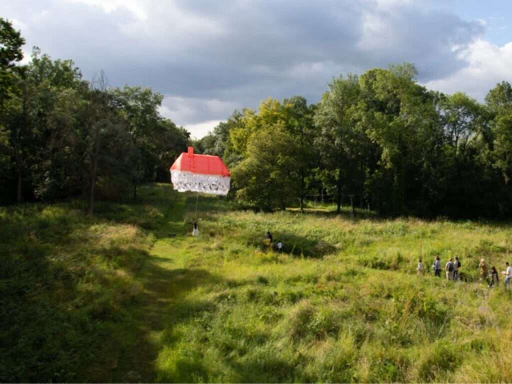 A wide landscape view showing a floating red and white house installation positioned over a vast green valley, with a small group of observers walking along a path on the right under a dramatic cloudy sky.