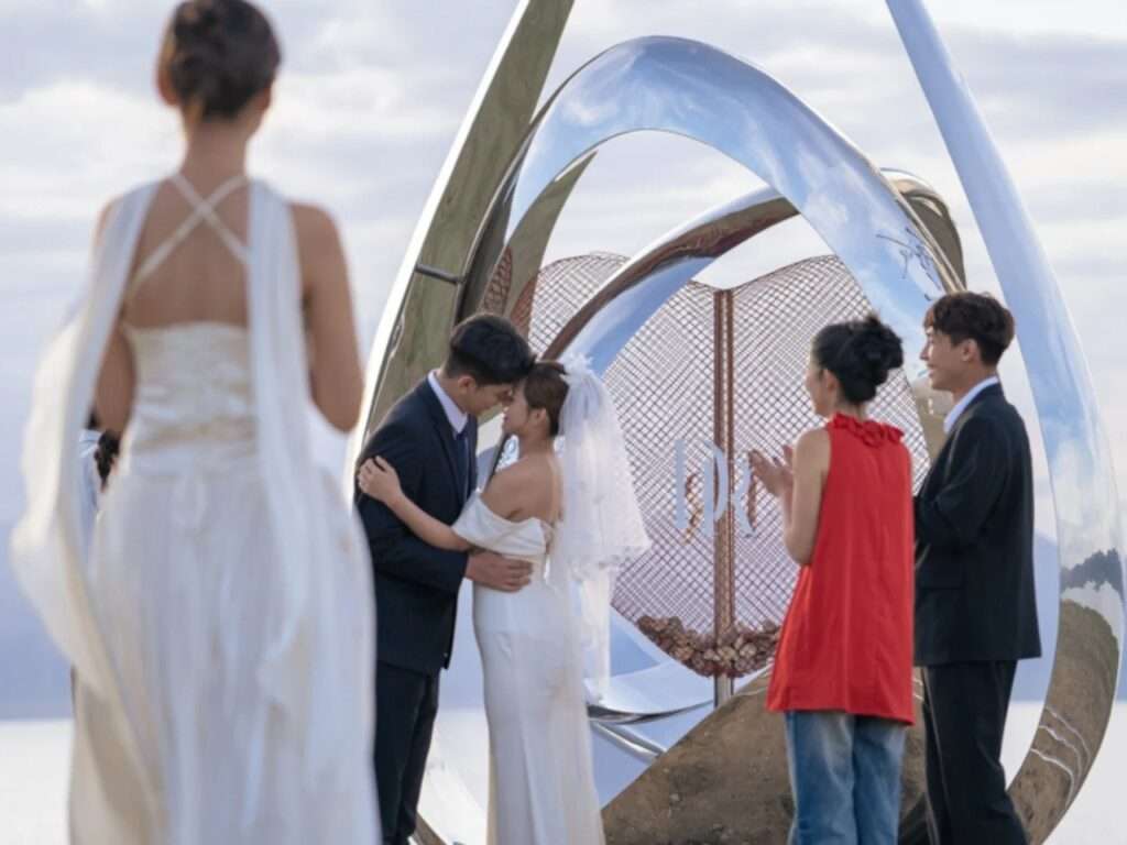 wedding couple embracing inside the frame of the reflective pavilion while guests watch and applaud during an outdoor ceremony.