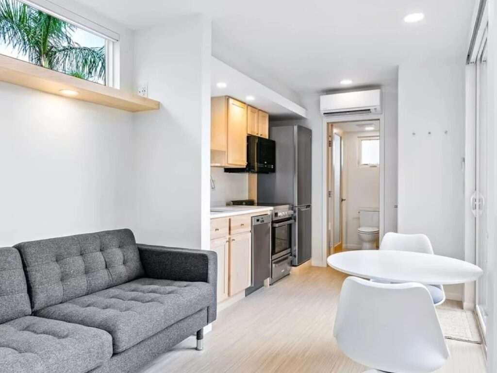 View from the living area of the N2 House showing the kitchen, an adjacent round dining table with two chairs, and the entrance to the bathroom.
