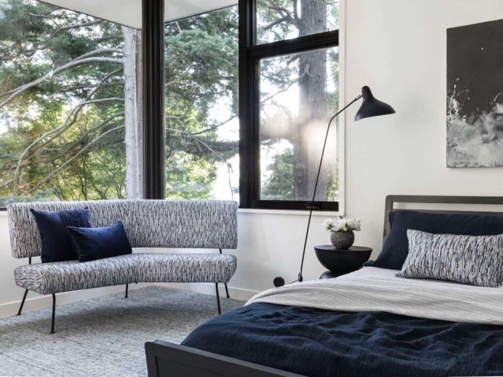 Minimalist master bedroom with a blue bedspread, a textured accent sofa, a black floor lamp, and large windows looking into the trees.