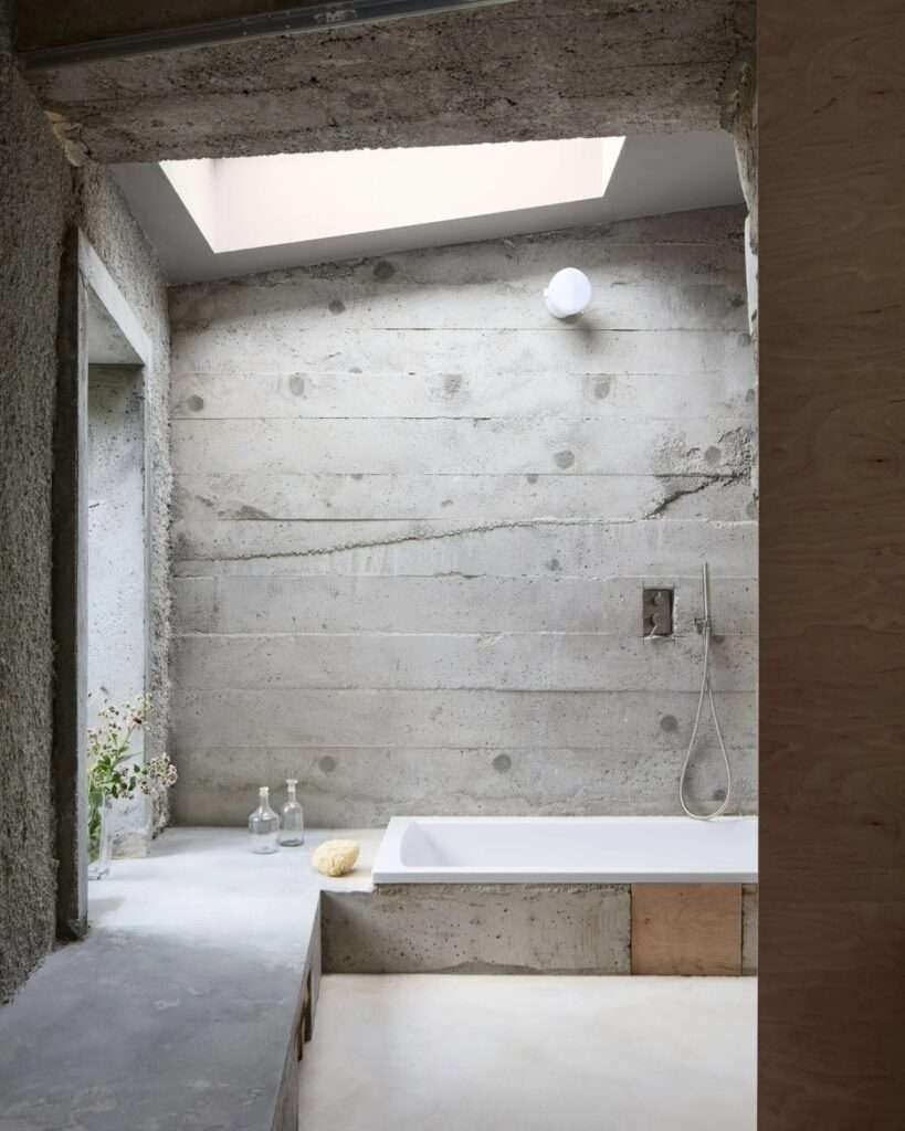 Sunken concrete bathtub beneath a skylight inside a minimalist bathroom with board-formed concrete and rough textured walls.