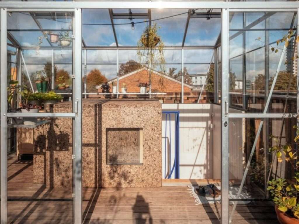 Glass greenhouse interior on the roof of Peckham House featuring a cork-clad sliding access hatch and blue metal handrail.