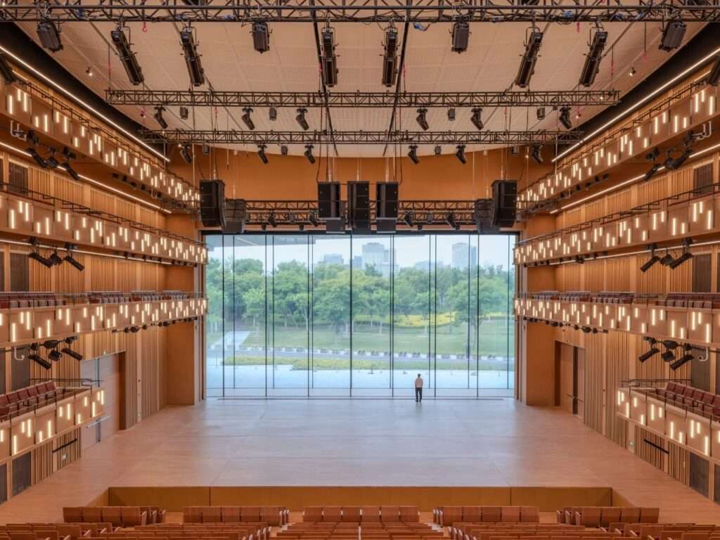 Interior view of an auditorium featuring dark wood textures, warm lighting, and a panoramic glass window looking out to greenery.
