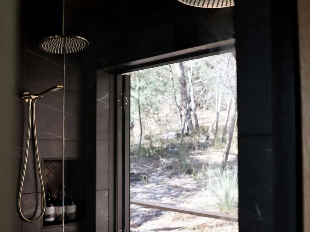 Detailed view of a modern cabin shower corner with dark marble-tiled walls, a copper-toned overhead rain shower, a hand shower, and large glass doors open to the forest view.