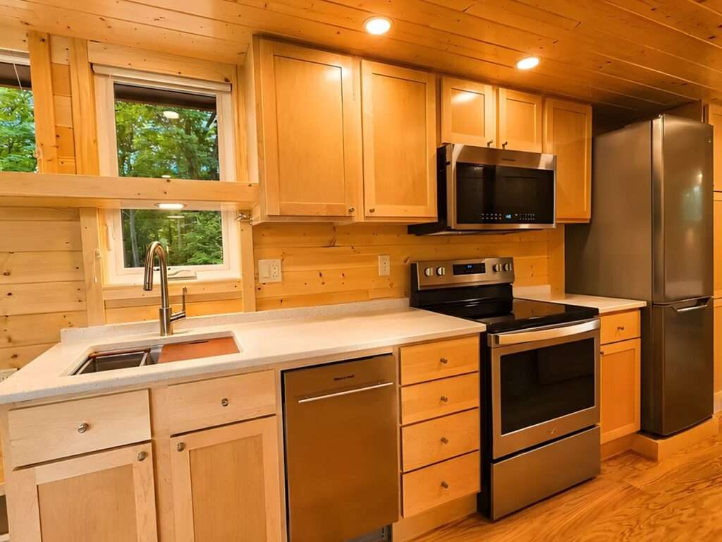 Close-up of a modern kitchen in a tiny home with wooden cabinets, a light countertop, a stainless steel sink, a dishwasher, a full-size range, and a refrigerator.