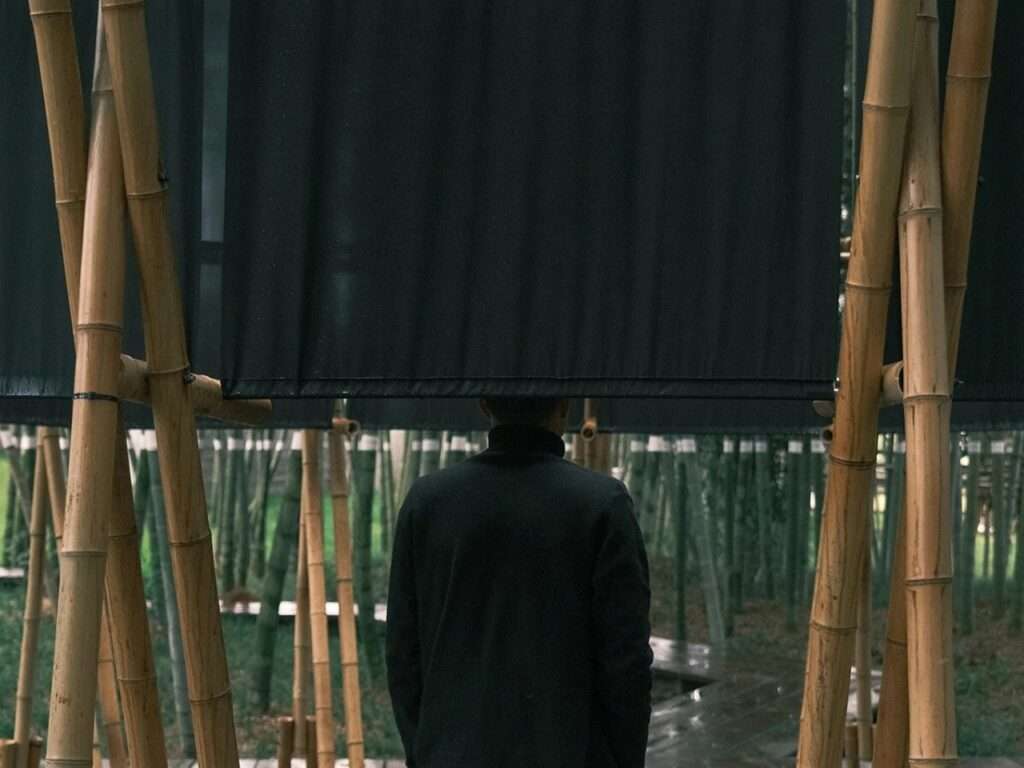 A person seen from behind facing a large, opaque black textile curtain suspended within a raw bamboo structural frame in a forest.