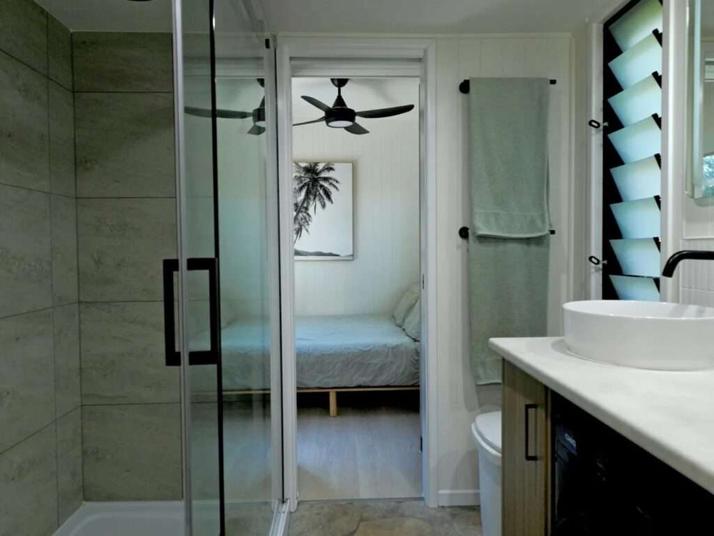 Bathroom interior view showing a glass shower enclosure with stone-look tiles, a louvre window, and a clear line of sight leading into the adjacent private bedroom.