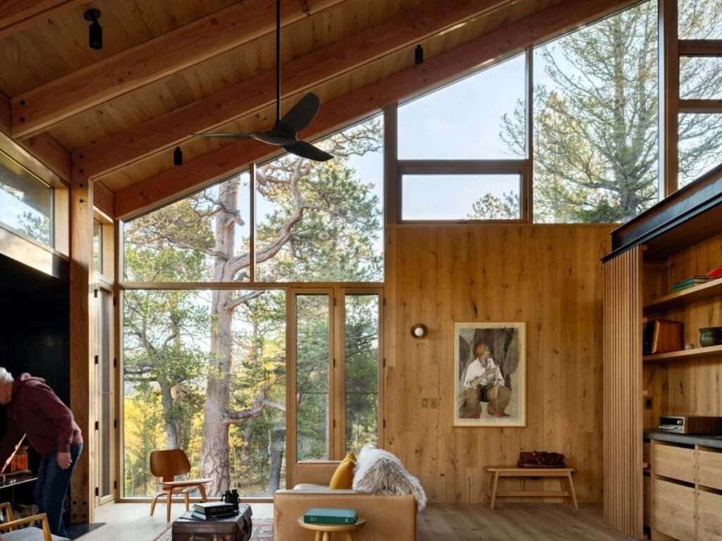 Spacious cabin living room with an open-plan layout, vaulted wood ceiling with a modern ceiling fan, large panoramic windows facing the forest, and warm oak wall paneling.