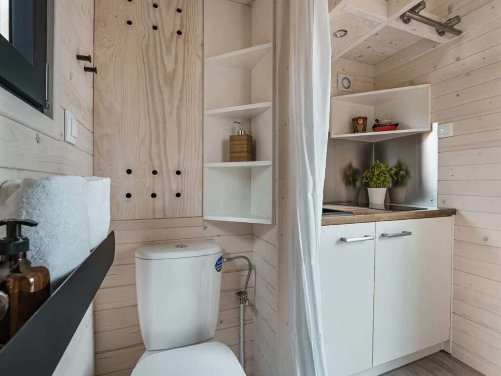 Tight angle interior of a micro bathroom with a white toilet, custom plywood shelving, and a fabric divider screen.