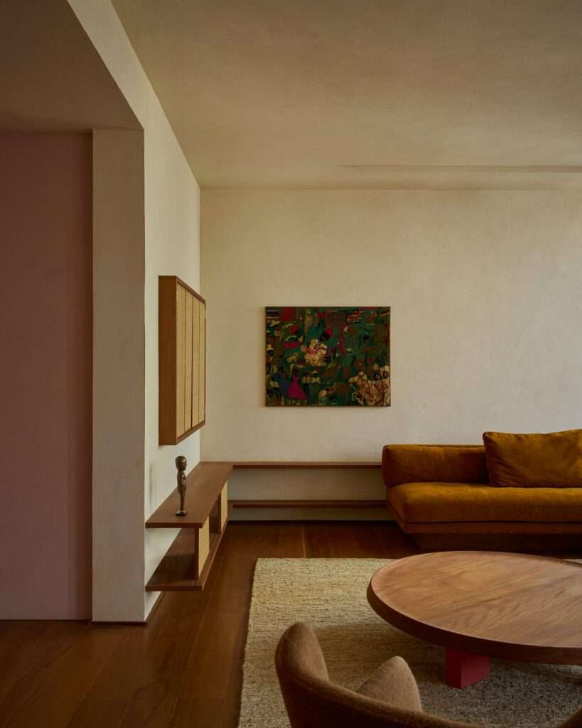 Detail of custom low floating wooden shelves mounted on a light textured wall, displayed alongside a framed woven artwork, a small sculpture, and the edge of a mustard velvet sofa.