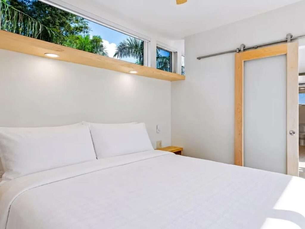 Modern loft bedroom in the N2 House, featuring a queen bed with white linens, a wooden sliding barn door, and a built-in wooden closet under high clerestory windows.