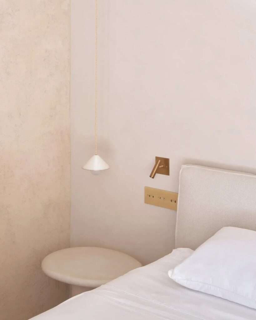 A close-up detail of a bedroom corner in Casa Óculo, showing a minimalist pendant light, a recessed stone nightstand, a textured wall finish, and a brass control panel.