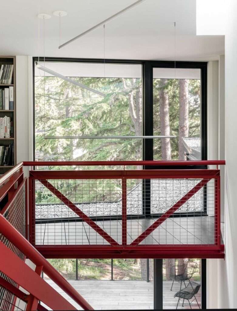 Architectural steel staircase railing painted in saturated red contrasting with white walls and looking out a large forest window.
