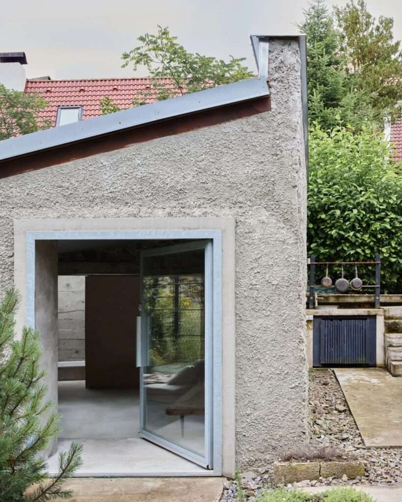 Exterior corner view of adaptive reuse house showing open glass door set into concrete frame and textured rough-cast wall.
