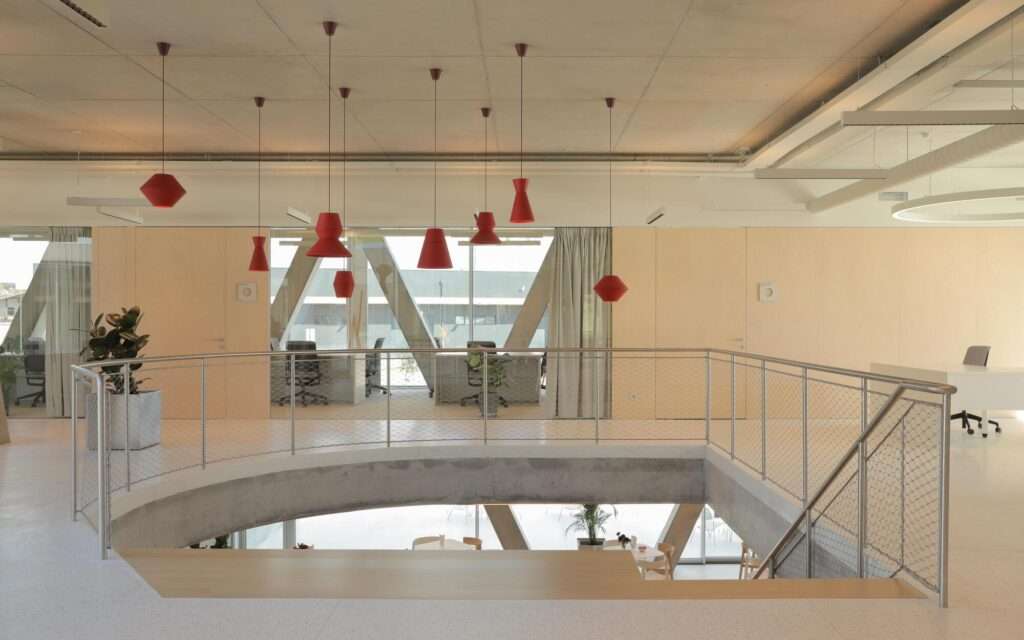 Interior atrium view looking down through a floor cutout with stainless steel wire mesh handrails and suspended decorative red lamps overhead.
