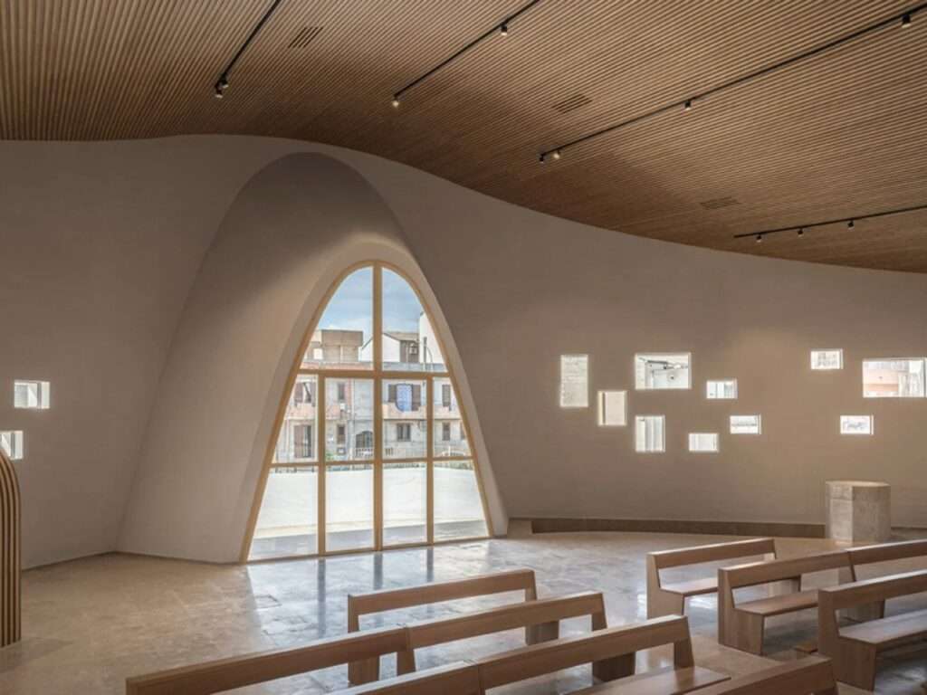Interior of the Santa Barbara Parish sanctuary showing wooden pews, a slatted timber ceiling, an arched window, and small square windows.