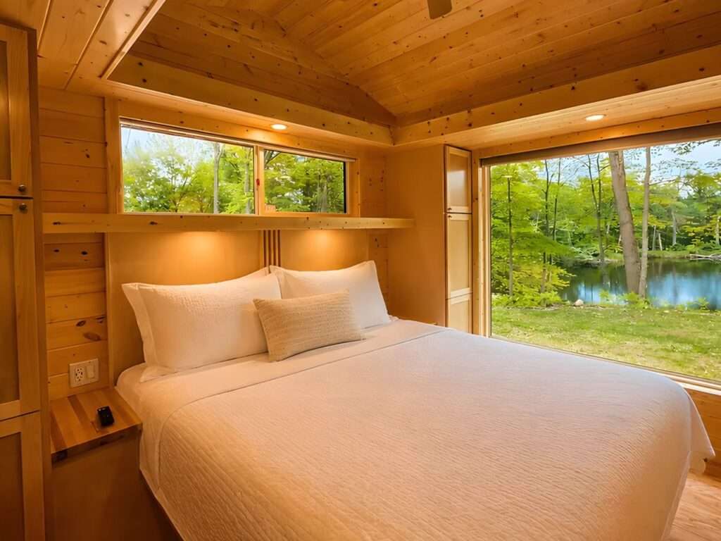 Cozy master bedroom with a king-size bed, white linens, all-wood walls, and a massive glass window framing a scenic view of a lake and green trees.
