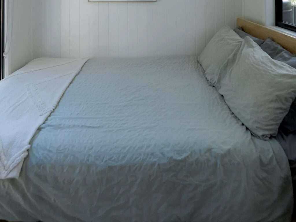 Minimalist master bedroom in the Burley 9.6 tiny house featuring a double bed with light grey bedding, white VJ vertical paneled walls, and a warm timber headboard.