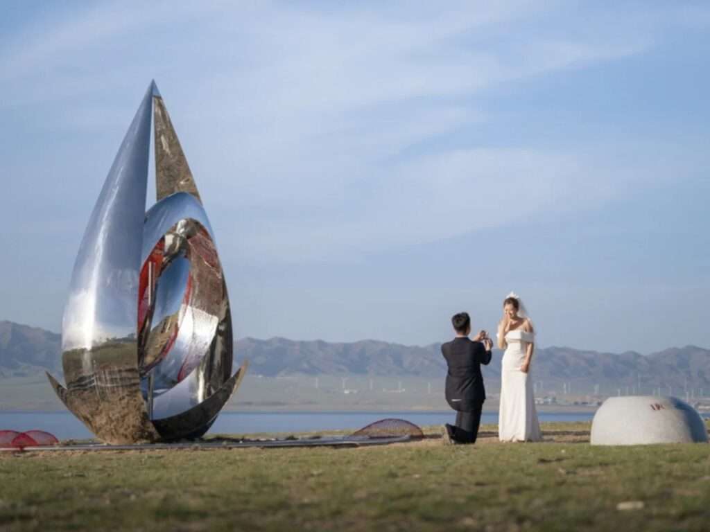 Side profile of the water-droplet chrome structure on a grassy plain with a man proposing to a bride in the background against a mountain range.