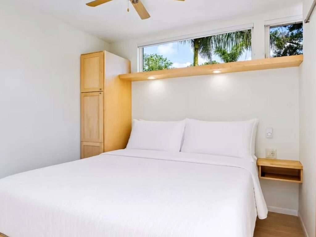 Panoramic view of the N2 House loft bedroom, showing a full queen bed with white linens, floating nightstands, clerestory windows with a palm tree view, and a ceiling fan.