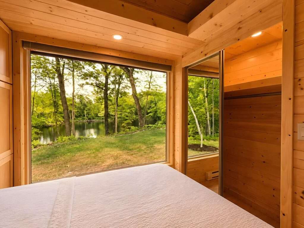A bedroom inside a wooden tiny home showcasing built-in mirrored wardrobes that reflect an external forest view through a large window.