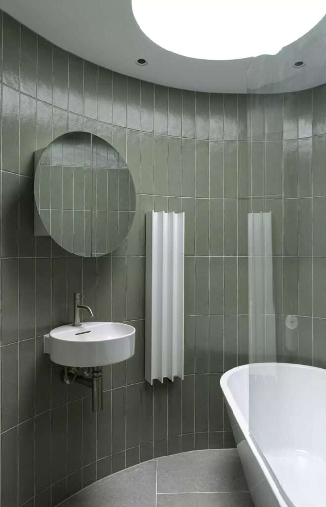 Curved bathroom wall lined with vertical olive green tiles featuring a white freestanding tub, floating sink, and circular skylight.