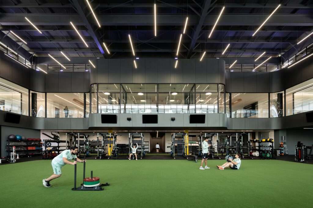 Double-height indoor training gym featuring green artificial turf flooring, strength training racks, linear ceiling lights, and an upper-level glass mezzanine administrative area.