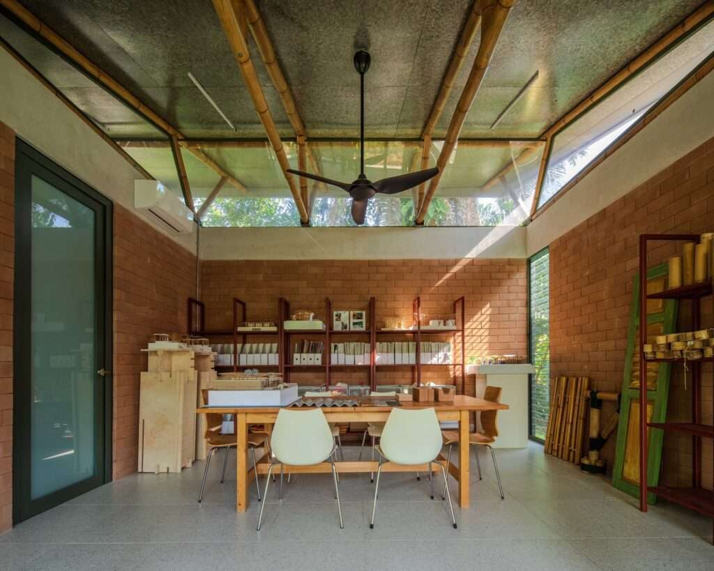 Interior meeting room featuring a wooden table, white modern chairs, compressed earth block walls, shelving units with bamboo models, and a ceiling fan under a high clerestory window framework.
