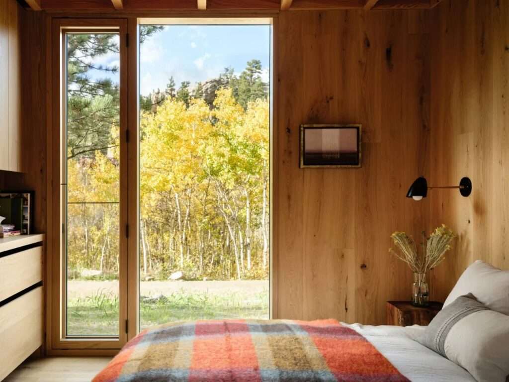 Minimalist cabin bedroom with oak walls, a low bed topped with a colorful plaid blanket, and a large floor-to-ceiling window framing bright yellow autumn foliage.