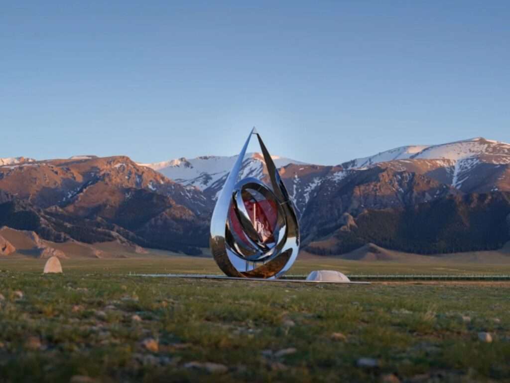 A wide view of the reflective teardrop pavilion sitting on a vast green meadow under a blue sky, with expansive snow-capped mountains spanning the background.