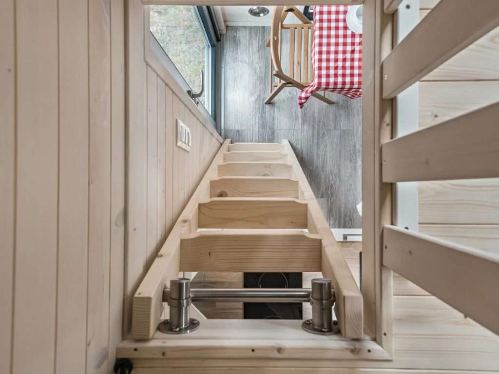 Looking down a light-colored wooden ladder inside La Ruche, connecting the upper loft bedroom to the ground floor below.