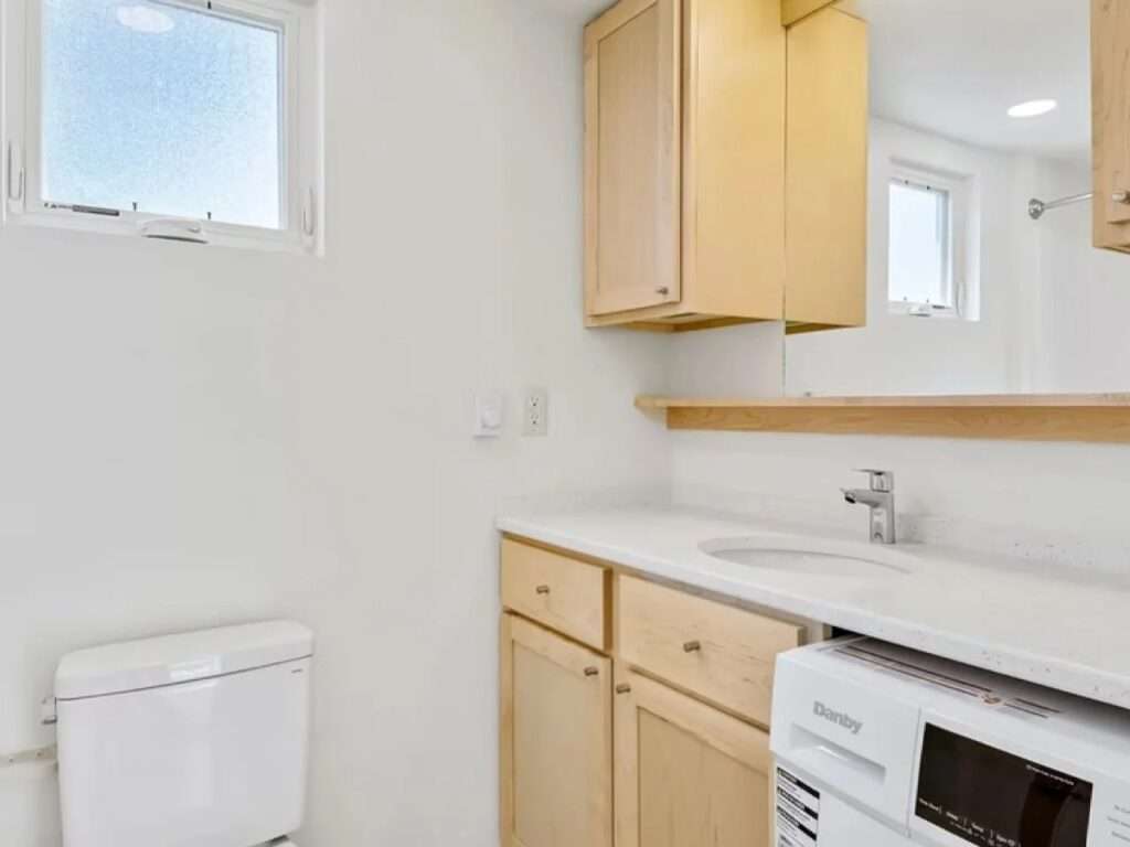 Compact but complete bathroom interior of the N2 House, with a light wood vanity, a full mirror, an integrated sink, a toilet, and an efficient washing machine.