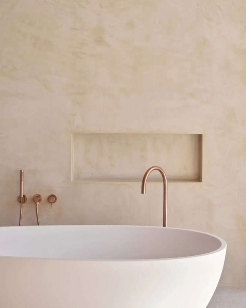 A close-up detail of a minimalist bathroom in Casa Óculo, focusing on a sculptural copper faucet and a white freestanding tub against a lime mortar wall with a recessed niche.